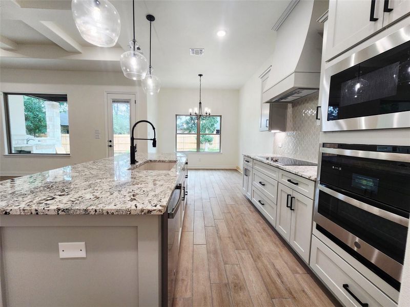 Open-concept kitchen featuring an expansive island with a built-in sink and modern faucet, granite countertops, wood-finish flooring, white cabinetry with dark hardware, and stainless steel built-in appliances