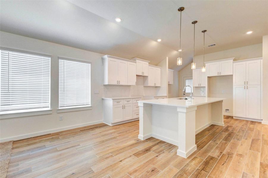 Modern kitchen with a large island, quartz countertops, white shaker-style cabinetry, pendant lights, stainless fixtures, and warm wood-look tile flooring. Two large windows bring in natural light and brighten the adjoining breakfast area.