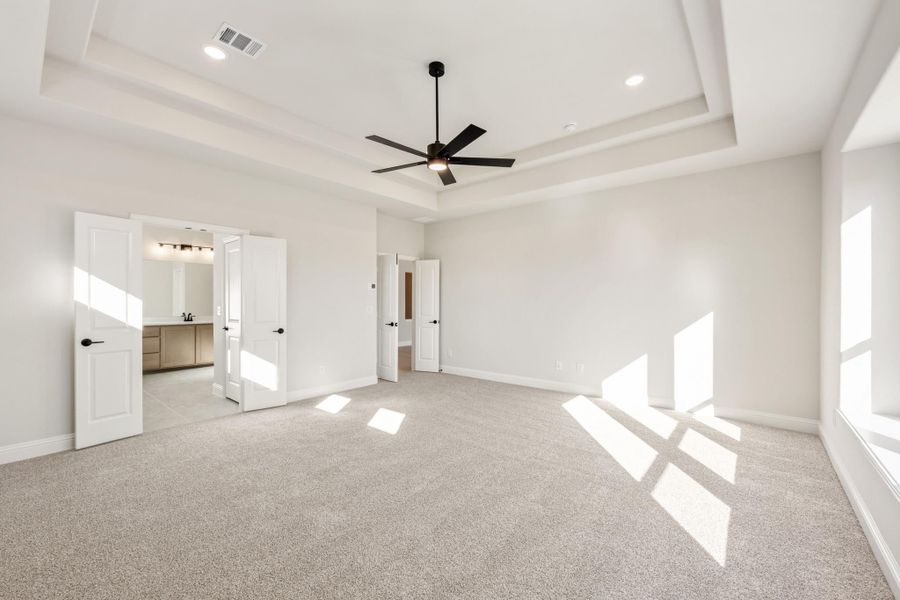Spacious bedroom with tray ceiling, ceiling fan, carpet flooring, and open door to ensuite bathroom