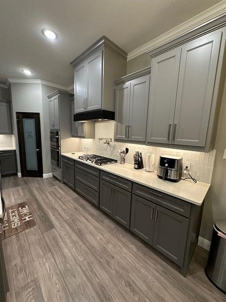 Kitchen with crown molding, decorative backsplash, gray cabinets, dark wood-style flooring, and light stone counters