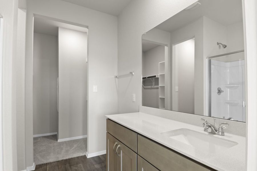 Image of a bathroom with a large mirror and white vanity with light brown cabinets with a walk-in shower reflected in the mirror. Open doorway to walk-in closet