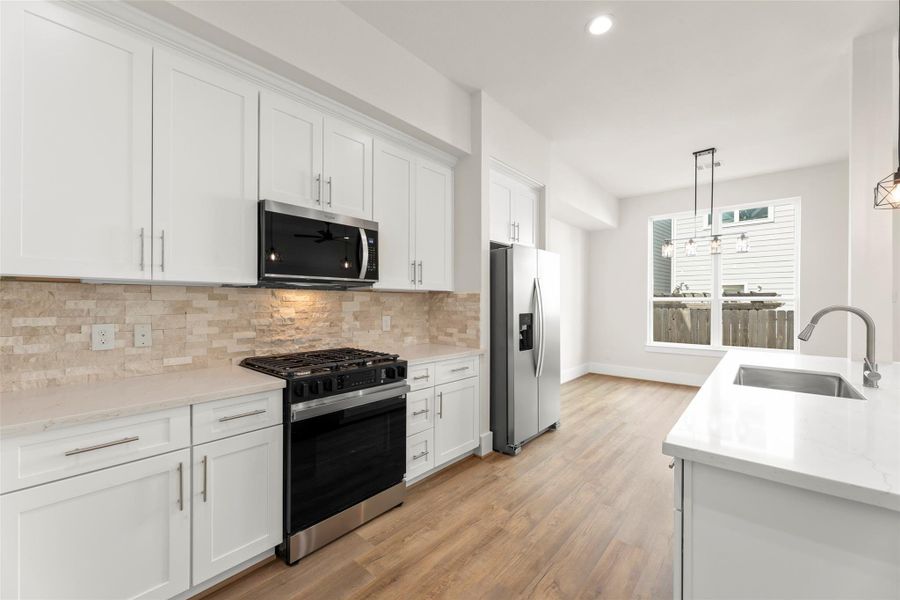 Modern kitchen with white cabinetry, stainless steel appliances, and a sleek island with sink. Light floors complement the neutral backsplash, and a large window provides natural light.
