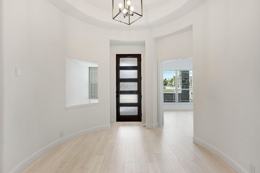 Rounded foyer with dark wood glass-panel front door, tray ceiling, pendant light, and light hardwood floors