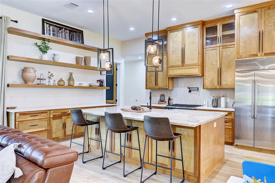 This modern kitchen features warm wood cabinetry, open shelving, and a large island with seating for three. It includes stainless steel appliances, pendant lighting, and ample counter space, creating a cozy and functional cooking area.