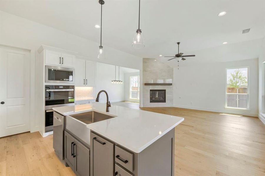 Kitchen island with a farmhouse sink and modern faucet, white cabinetry, built-in stainless steel appliances, wood-finish flooring, and a fireplace with a stone surround Kitchen island with a farmhouse sink and modern faucet, white cabinetry, built-in stainless steel appliances, wood-finish flooring, and a fireplace with a stone surround