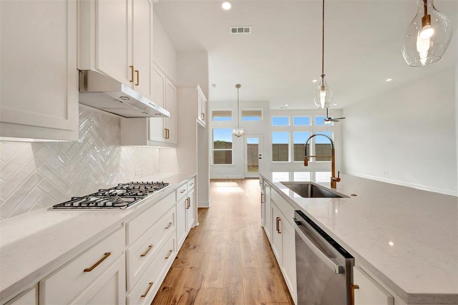 Open-concept kitchen featuring an expansive island with an undermount sink and contemporary faucet, wood-finish flooring, white cabinetry with gold-tone hardware, a herringbone pattern tile backsplash, and a gas cooktop with a range hood