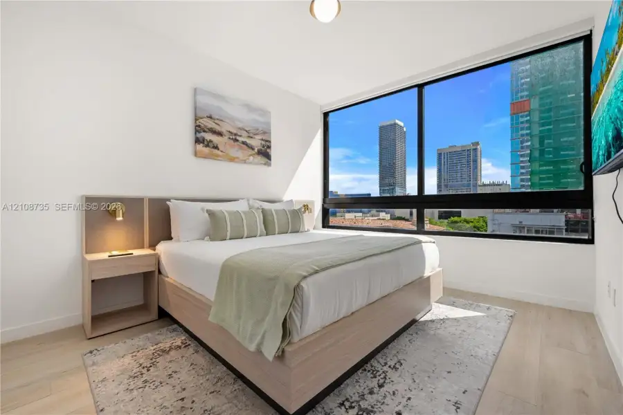 Floor-to-ceiling windows featuring urban scenery, built-in wooden bed frame with integrated headboard, and light wood-finish flooring Floor-to-ceiling windows featuring urban scenery, built-in wooden bed frame with integrated headboard, and light wood-finish flooring