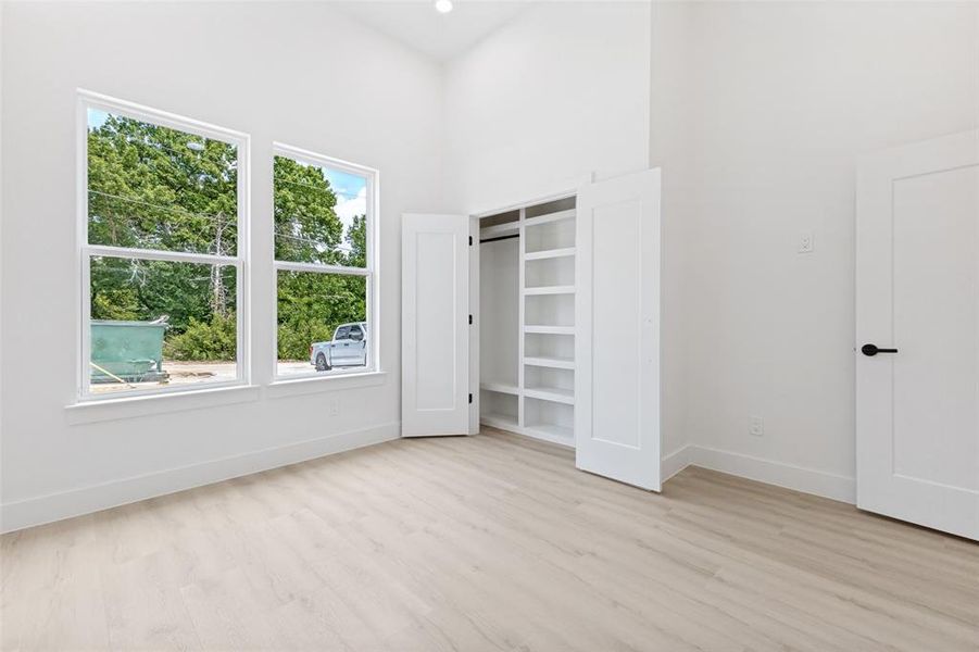 Room featuring high vaulted ceilings, large double-hung windows, wood-finish flooring, a built-in closet system with shelving, and a modern interior door with matte black hardware Room featuring high vaulted ceilings, large double-hung windows, wood-finish flooring, a built-in closet system with shelving, and a modern interior door with matte black hardware