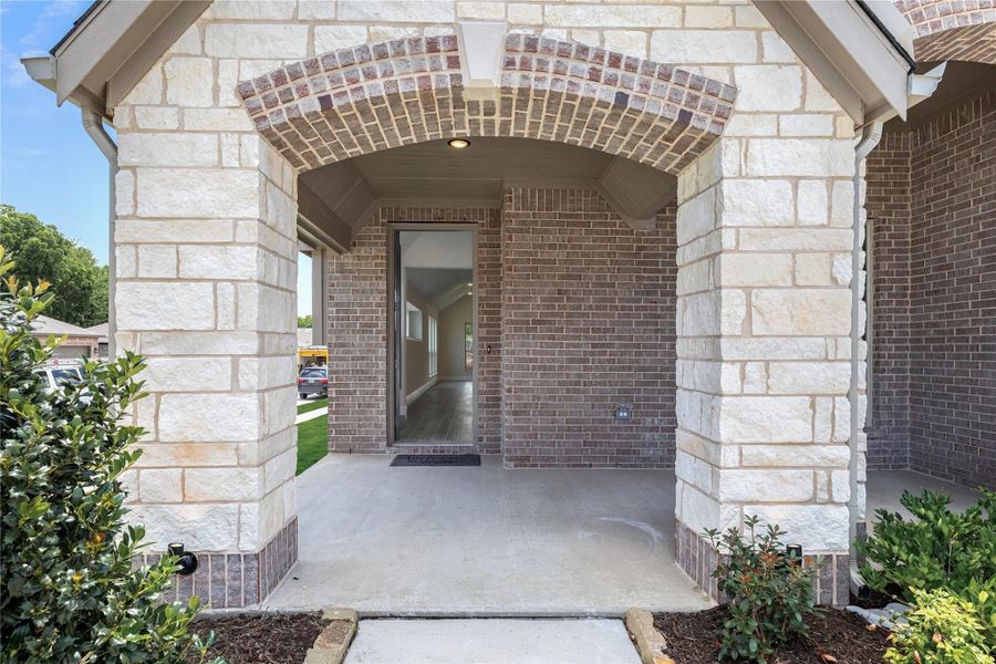 Covered front entry with brick and stone archway, open front door, and concrete porch with landscaping Covered front entry with brick and stone archway, open front door, and concrete porch with landscaping