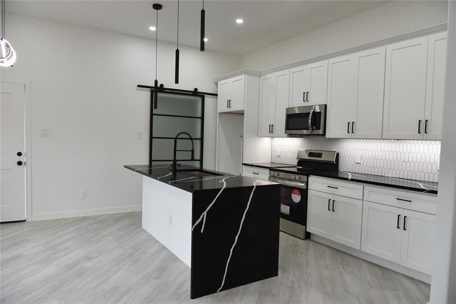 Kitchen featuring hanging light fixtures, stainless steel appliances, a barn door, a center island with sink, and white cabinets