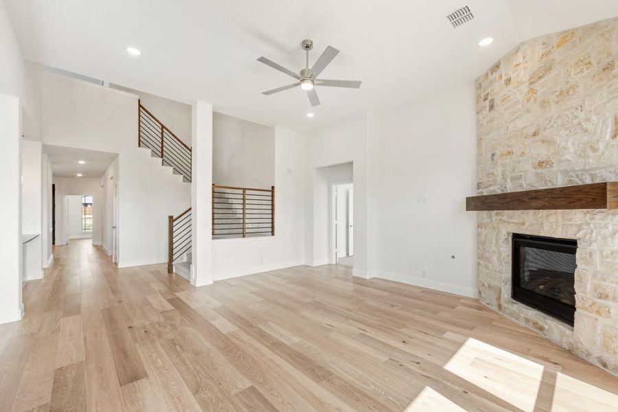 Open living room with hardwood floors, stone fireplace, ceiling fan, and staircase with metal railing Open living room with hardwood floors, stone fireplace, ceiling fan, and staircase with metal railing