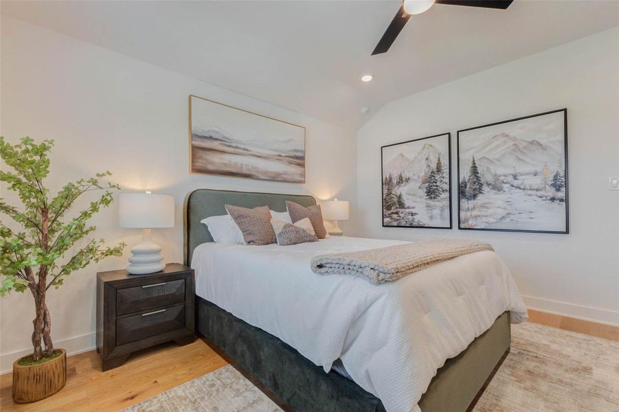 Soft textures and calming tones give this bedroom a warm, relaxing character. The upholstered bed is framed by matching nightstands and sculptural lamps, while oversized landscape artwork introduces a peaceful natural theme. Warm wood floors, a textured area rug, recessed lighting, and a contemporary ceiling fan complete the comfortable and cohesive design.