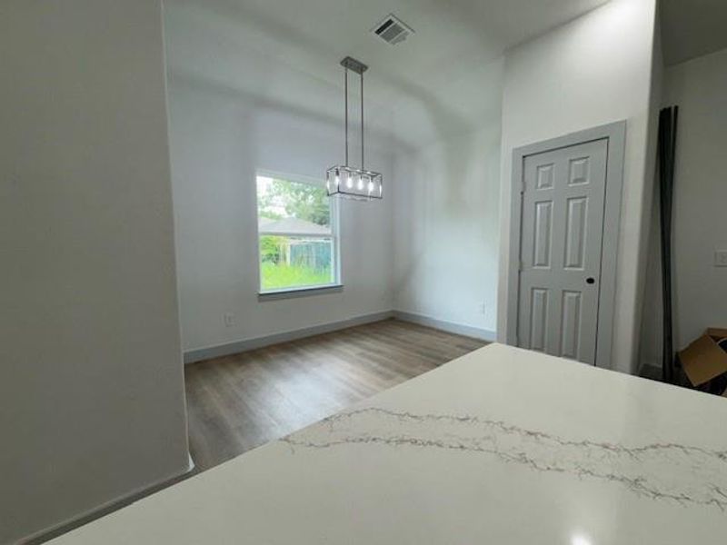 This is a modern, bright dining space featuring a large window for natural light, high ceilings, elegant pendant lighting, and sleek laminate flooring. The view suggests a clean, open-plan design with a glimpse of a stylish countertop in the foreground, indicating a contemporary kitchen adjacent to the dining area.