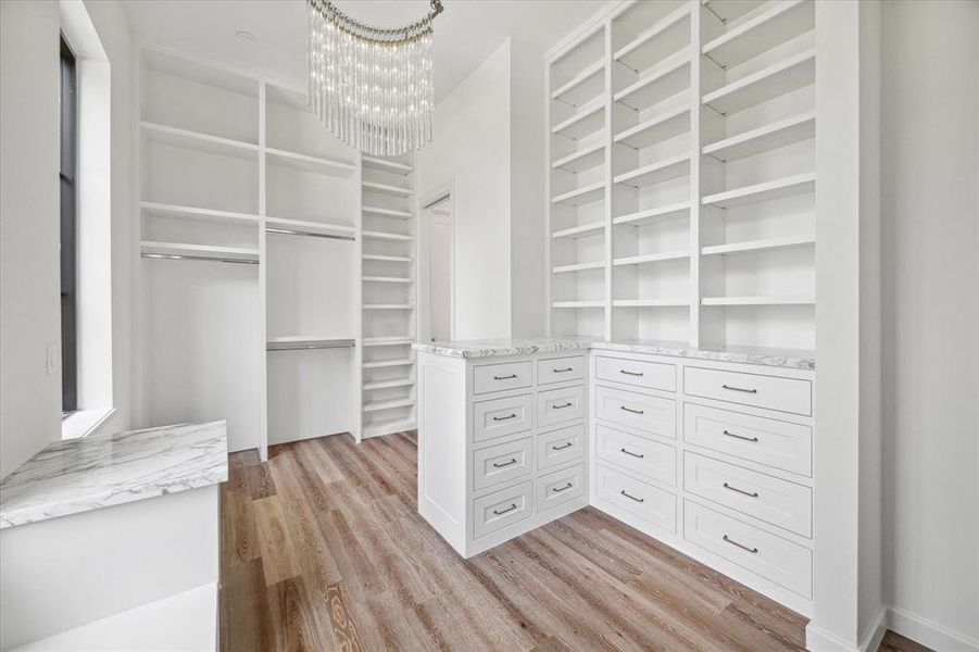 This luxurious walk-in closet features custom built-in shelving and inset cabinetry with marble countertops. The ample storage space, including drawers and open shelves, ensures organization and elegance. A large window allows natural light to fill the room, highlighting the sophisticated chandelier that adds a touch of glamor to the space. The warm hardwood floors complete the inviting and functional design.