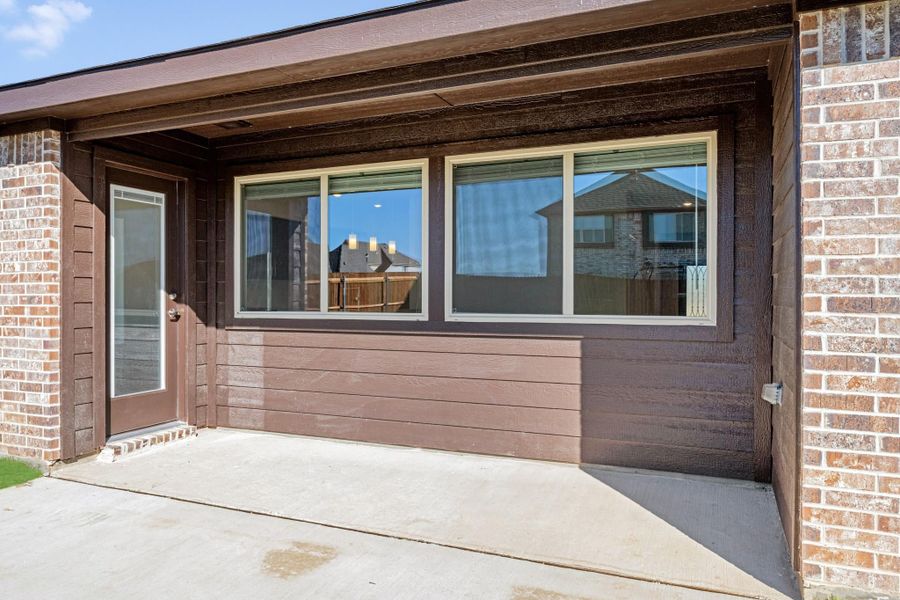 Covered concrete patio with brick exterior, brown siding, large windows, and a side entry door