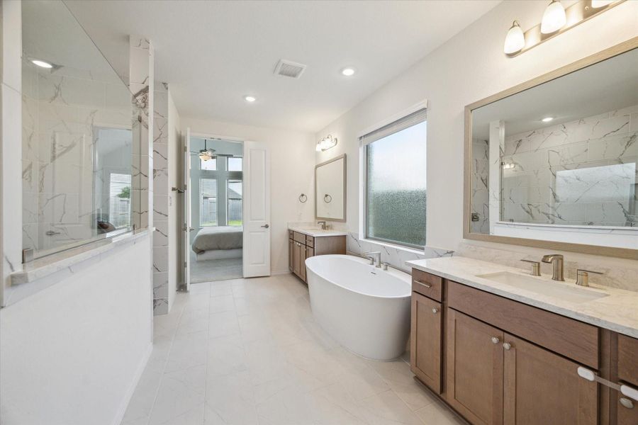 Full perspective of the primary bath highlighting all features, including double vanities, soaking tub, and floor-to-ceiling tiled shower looking back into the primary bedroom.