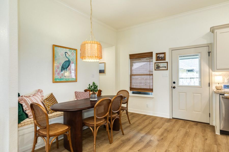 This cozy dining area features a built-in bench with cushions, a wooden table, and chairs. Warm, natural light fills the space through a window with bamboo style blinds. A stylish pendant light hangs overhead, and the area is accented with artwork and neutral walls, providing a welcoming atmosphere.