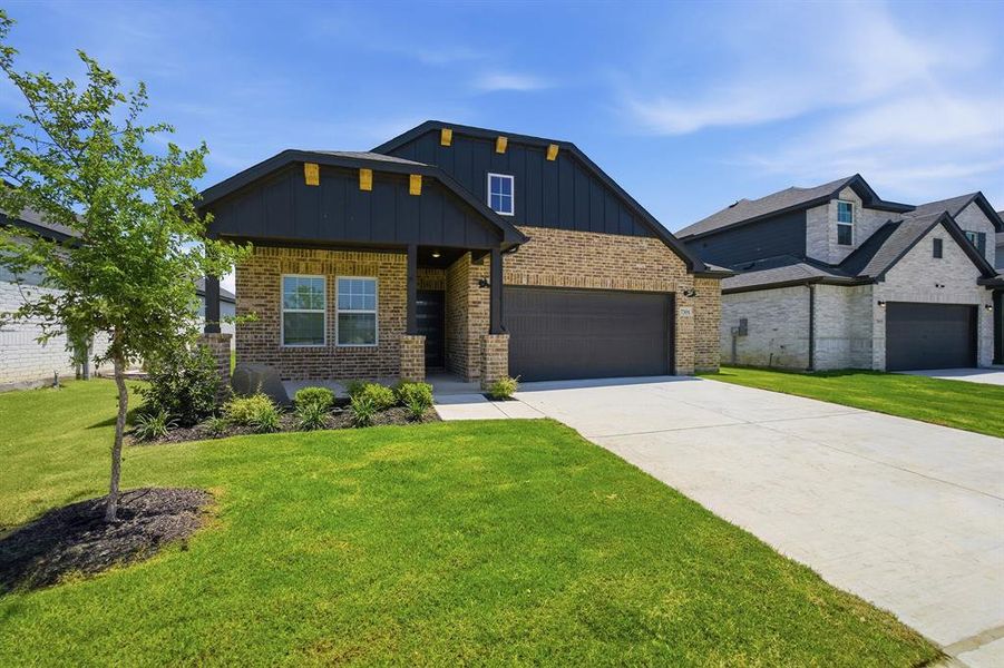 Contemporary residence featuring a brick and dark siding exterior, an attached garage, a covered front porch, a concrete driveway, and a landscaped front yard with a young tree