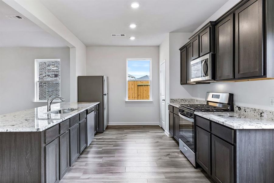 Kitchen with appliances with stainless steel finishes, sink, dark brown cabinetry, light wood-type flooring, and light stone countertops