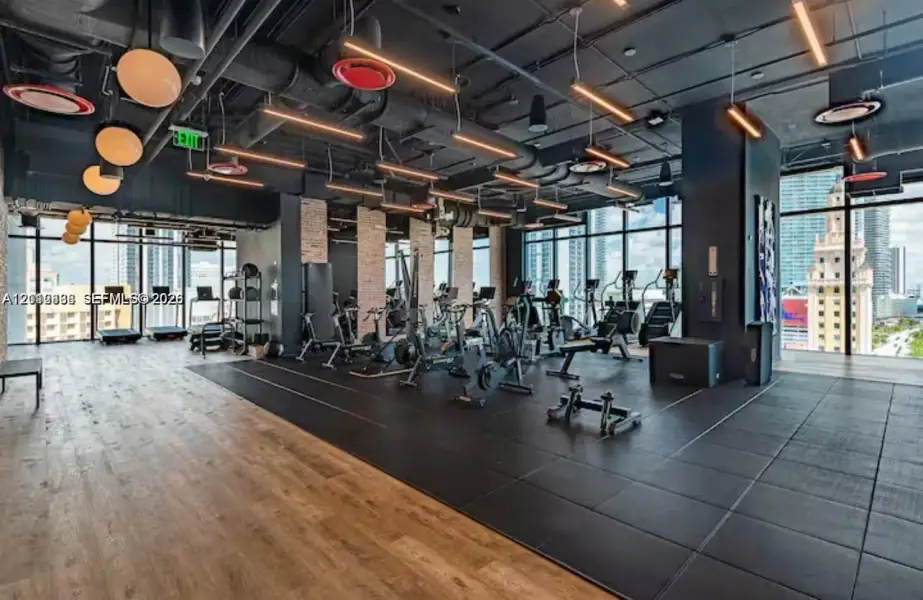 Industrial-style fitness center featuring floor-to-ceiling windows, exposed structural ceiling, wood-finish flooring, rubberized workout surfacing, and decorative globe lighting Industrial-style fitness center featuring floor-to-ceiling windows, exposed structural ceiling, wood-finish flooring, rubberized workout surfacing, and decorative globe lighting