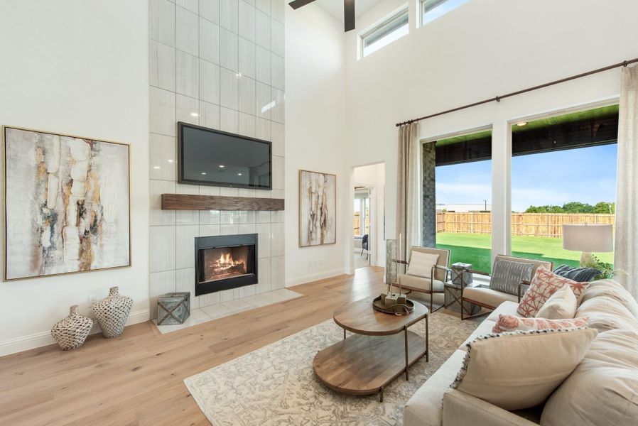 Living room with two-story ceiling, tiled fireplace, wall-mounted TV, hardwood floors, and large windows overlooking backyard Living room with two-story ceiling, tiled fireplace, wall-mounted TV, hardwood floors, and large windows overlooking backyard