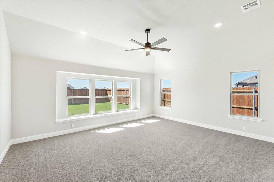 Spacious empty room with gray carpet, ceiling fan, and large windows overlooking a fenced backyard Spacious empty room with gray carpet, ceiling fan, and large windows overlooking a fenced backyard