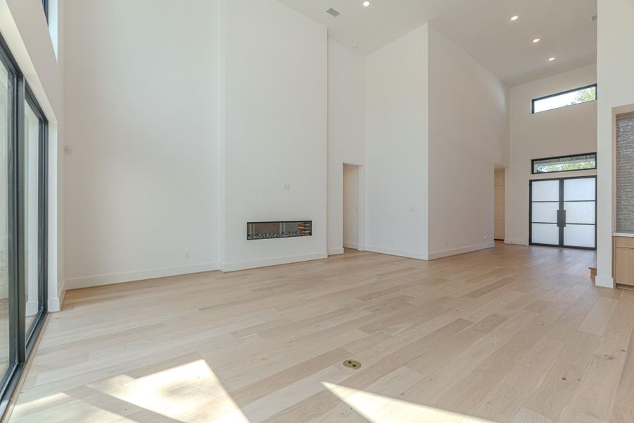 This spacious, modern living area features high ceilings, abundant natural light from large windows, and sleek hardwood floors. It includes a minimalist built-in fireplace and a stylish entryway with frosted glass doors.