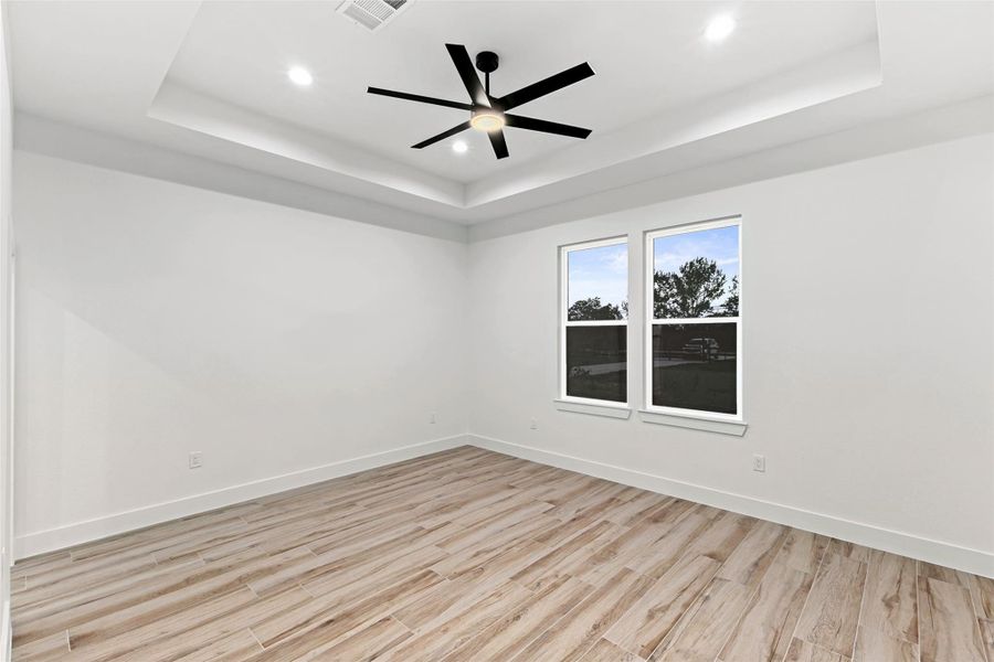 This primary bedroom features a modern design with light wood-look flooring, a tray ceiling with recessed lighting, and a sleek ceiling fan. Two large windows provide ample natural light. This primary bedroom features a modern design with light wood-look flooring, a tray ceiling with recessed lighting, and a sleek ceiling fan. Two large windows provide ample natural light.