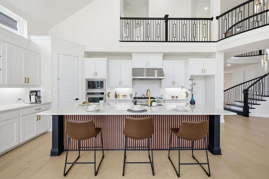 Gourmet kitchen featuring a central island with paneling, white cabinetry, integrated stainless steel appliances, light wood-finish flooring, and a two-story ceiling with an overlooking balcony