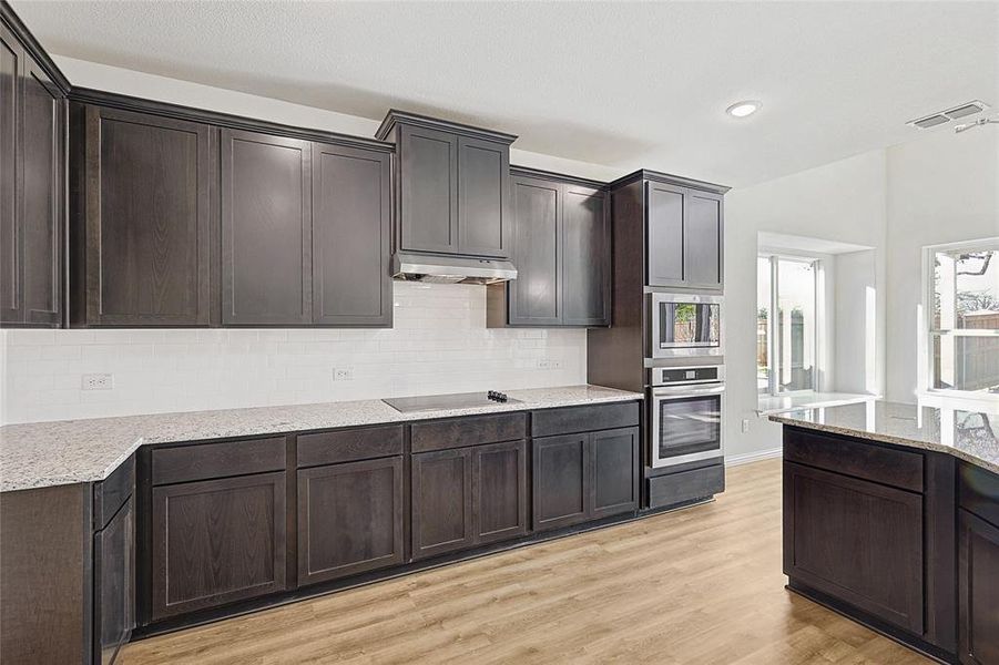 Kitchen featuring light hardwood / wood-style flooring, decorative backsplash, appliances with stainless steel finishes, dark brown cabinets, and light stone counters