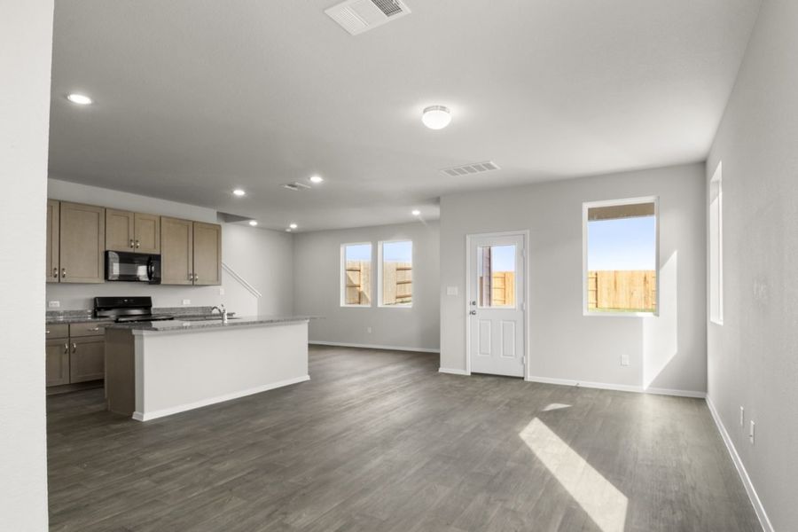 Image of a living room with light grey walls, dark brown floors, windows and a kitchen in the distance