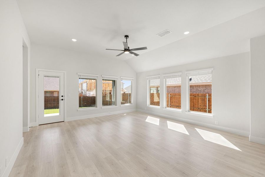 Bright open living room with vaulted ceiling, ceiling fan, light wood floors, and large windows overlooking backyard.