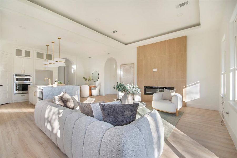 Living area featuring a tray ceiling, a large fireplace, light wood finished floors, and arched walkways Living area featuring a tray ceiling, a large fireplace, light wood finished floors, and arched walkways