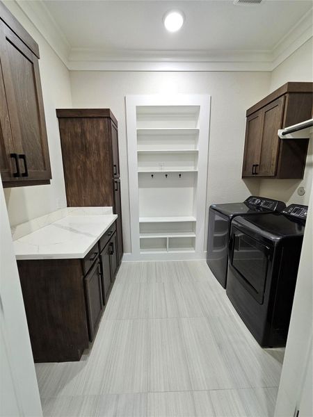 This photo shows a modern laundry room featuring a washer and dryer, wooden cabinets for storage, and a countertop with a marble-like surface. The room is well-lit with recessed lighting and has a built-in shelving unit.