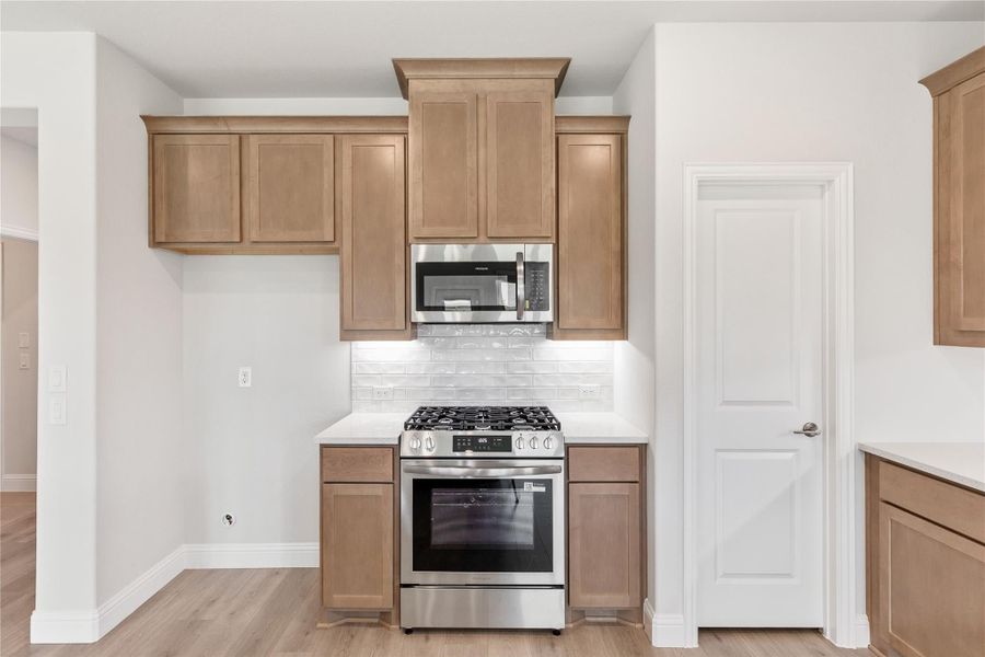 Kitchen with light wood cabinets, stainless steel gas range, microwave, and white tile backsplash