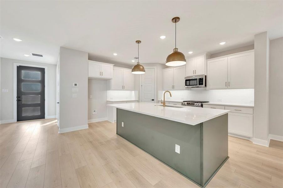 Two tone kitchen with an island with sink, hanging light fixtures, stainless steel appliances, light wood-style flooring, and light stone countertops