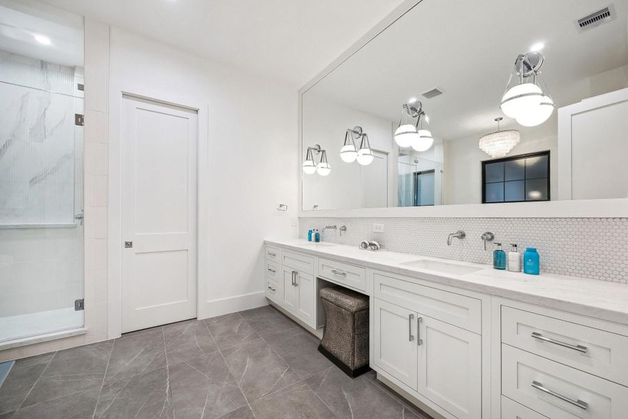 Primary Bath with 15 x 30 Matt Glazed Tile Floor with Subtle Veining.  Inset Shaker Style Cabinetry with Soft Close Drawers and Doors. Long Double Vanity with Slab Quartz Counter and a Penny Round Mosaic Tile Backsplash.  Dual Rectangle Sinks, Wall Mounted Faucets in Polished Chrome, Frameless Mirror with Four Globe Style Pendent Lights.  There is also a knee space and Separate Commode Area.
