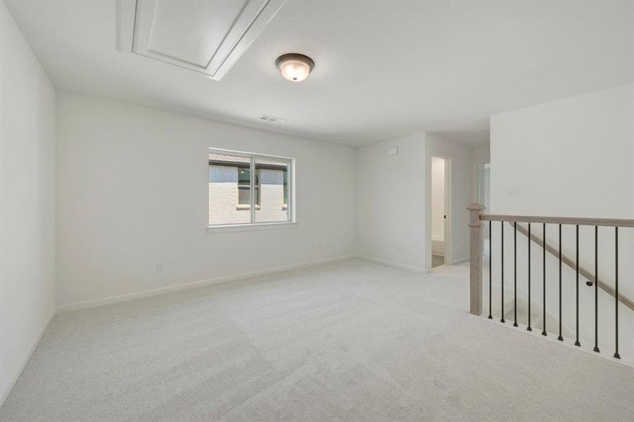 Carpeted upper-level landing featuring a large window, a ceiling light fixture, a pull-down attic access panel, and a wood and wrought iron staircase railing