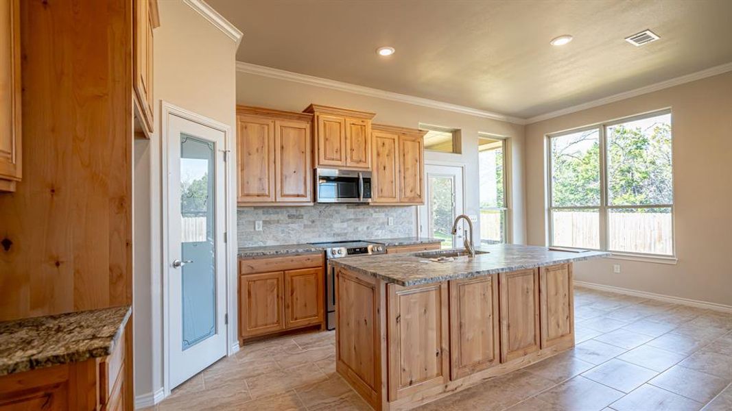 Kitchen with light stone counters, stainless steel appliances, tasteful backsplash, a kitchen island with sink, and crown molding