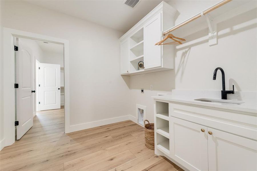 Dedicated laundry room featuring light wood-finish flooring, white cabinetry with gold-tone hardware, a white countertop with an undermount sink and matte black faucet, and a clothes hanging rod