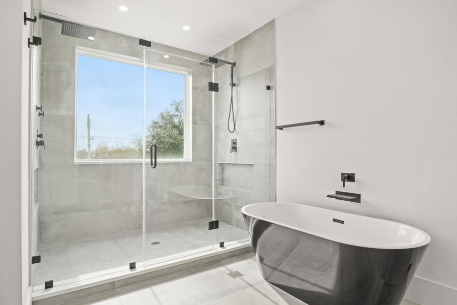 This view of the bathroom highlights the modern finishes between the freestanding tub with waterfall faucet and wide glass enclosed shower with recessed lights, fixed window, two overhead rain shower heads, one with a wand, the other with four additional body sprays, hexagonal tile floor and a large shampoo and soap niche.