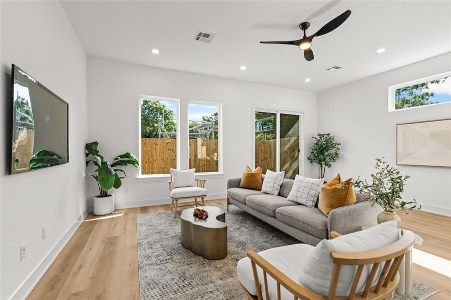 The main living room is bright and open, featuring a modern matte black ceiling fan and light oak flooring. Large windows look out into the backyard, providing a clean view of the horizontal wood fence and greenery while letting in plenty of natural light.