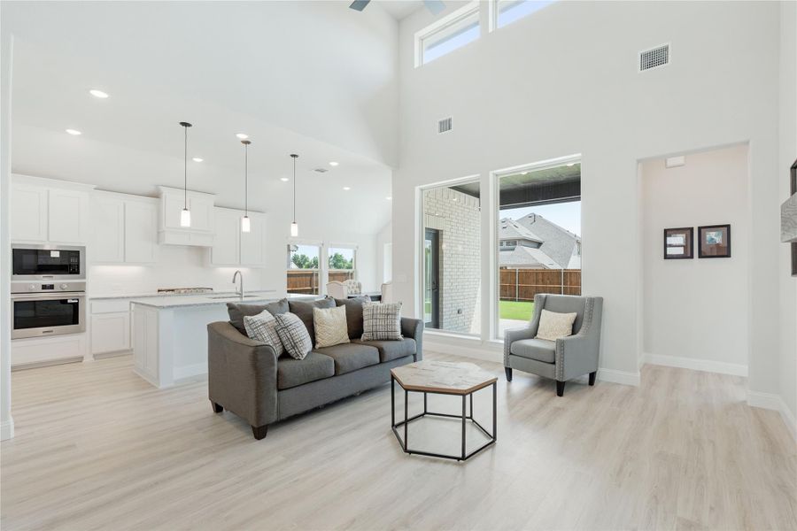 Open-concept living room with vaulted ceiling, light wood floors, and white kitchen visible in background