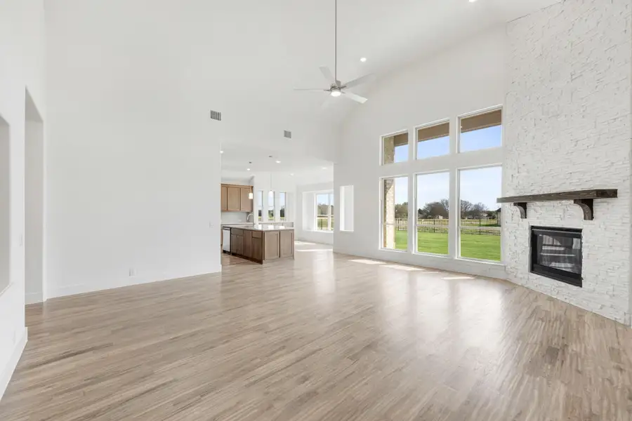 Open living room with vaulted ceiling, white brick fireplace, wood floors, and open kitchen visible in background. Open living room with vaulted ceiling, white brick fireplace, wood floors, and open kitchen visible in background.