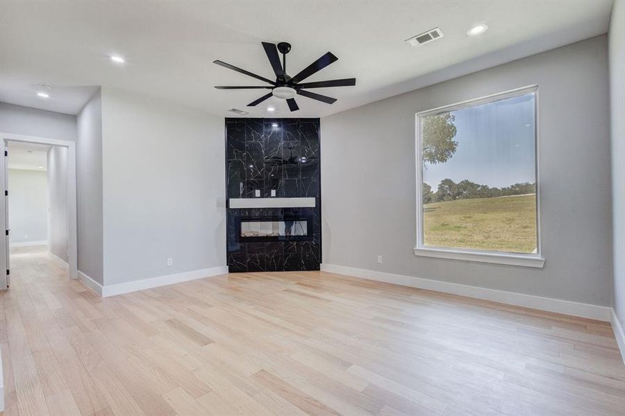 Spacious room featuring light wood-finish flooring, a contemporary black marbled fireplace surround with a white mantel, and a multi-blade ceiling fan