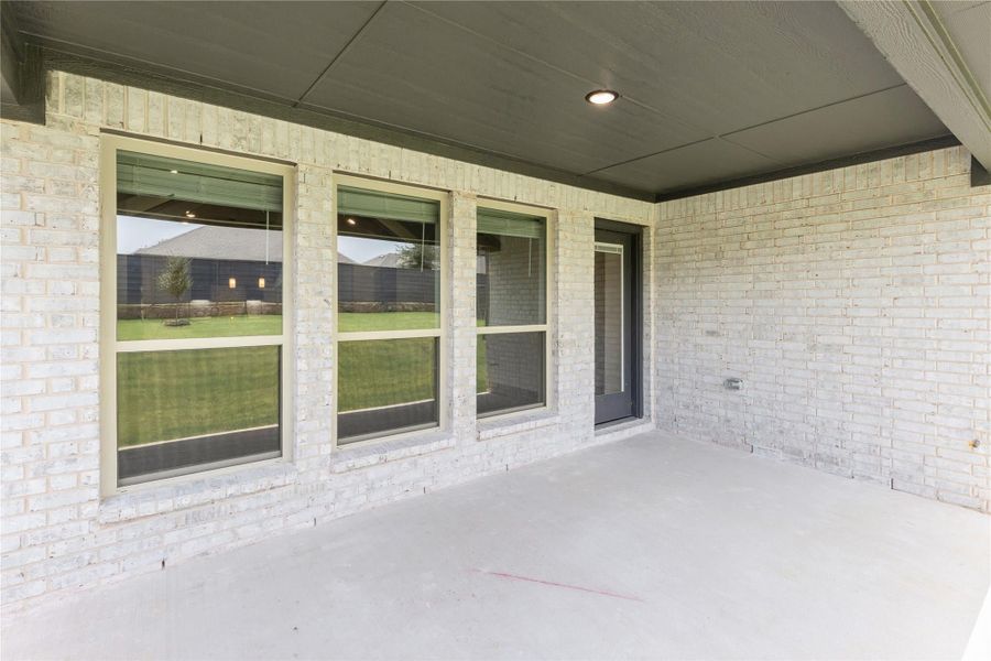 Covered back patio with concrete floor, light brick walls, three large windows, and a back door overlooking a grassy yard. Covered back patio with concrete floor, light brick walls, three large windows, and a back door overlooking a grassy yard.