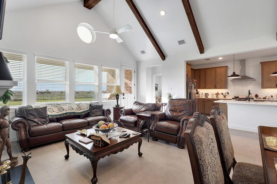 This inviting living space features high ceilings with exposed wooden beams, abundant natural light from large windows, and an open-concept layout connecting to a modern kitchen with a plethora of cabinet space.
