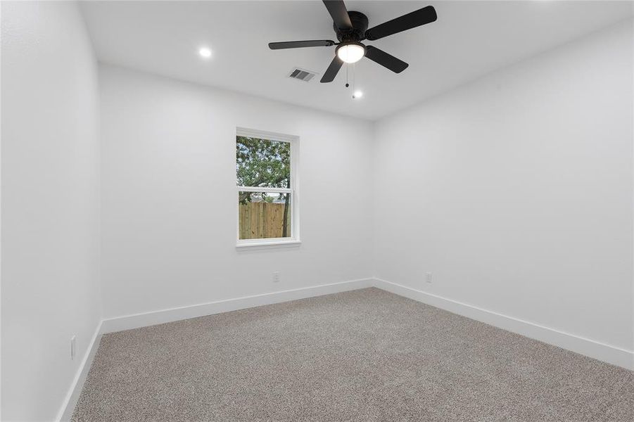 This is a bright, empty room with neutral walls and carpet flooring, featuring a single window and a ceiling fan with a light fixture.