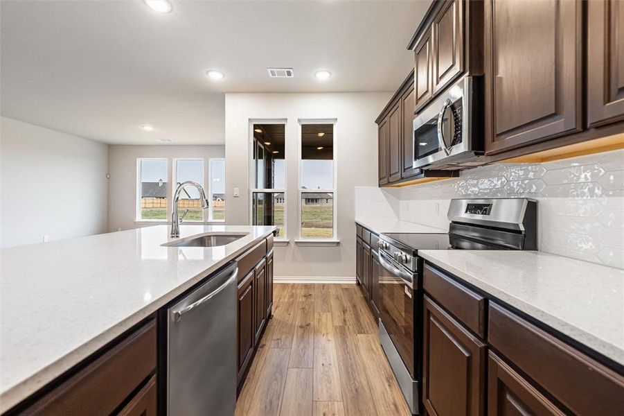 Kitchen featuring stainless steel appliances, dark brown cabinets, light stone countertops, light wood finished floors, and tasteful backsplash