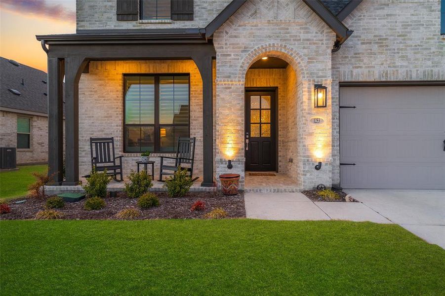 Exterior entry at dusk with a porch, an attached garage, central air condition unit, brick siding, and a yard